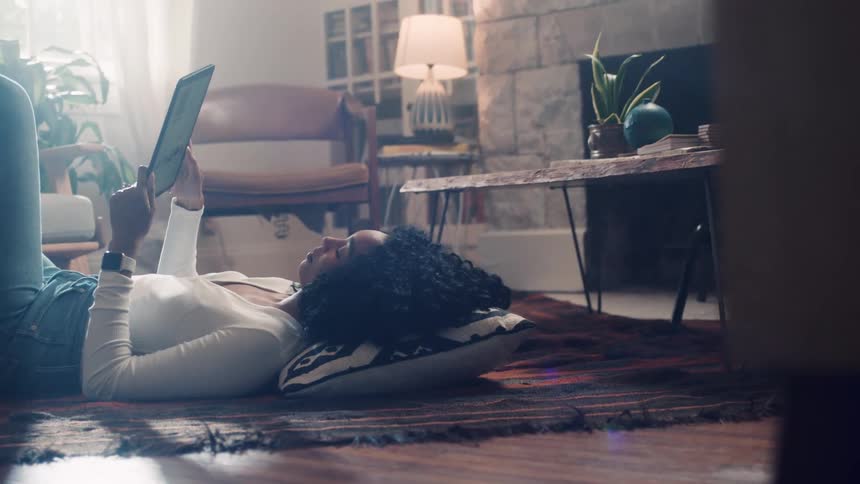 Black woman lying down on the floor uses a tablet in the living room at home