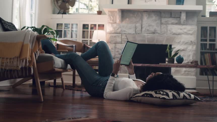 A woman laying on the floor with her head on a pillow and her foot on a couch while watching a tablet