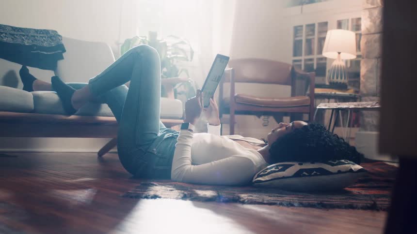Black woman lying down on the ground swipes down on her tablet in the living room at home