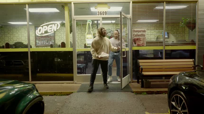 Two men walk out of a small diner with their food and walk to their car