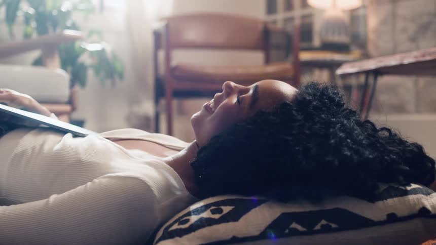 Black woman lying down on the floor holds a tablet and laughs in the living room at home