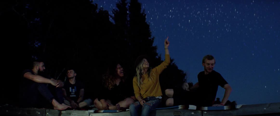 A group of friends sitting on the ground looks up at stars and laugh