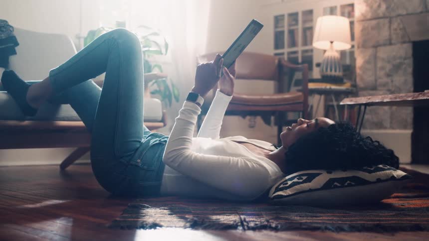 A woman laying on the floor with her head on a pillow and a foot on the couch using a tablet