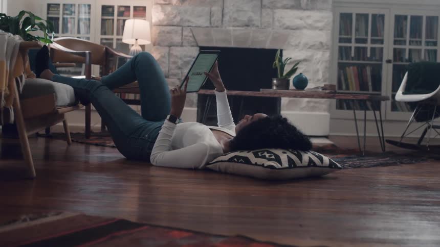 A woman uses a tablet while laying on the floor in her living room in front of a fireplace