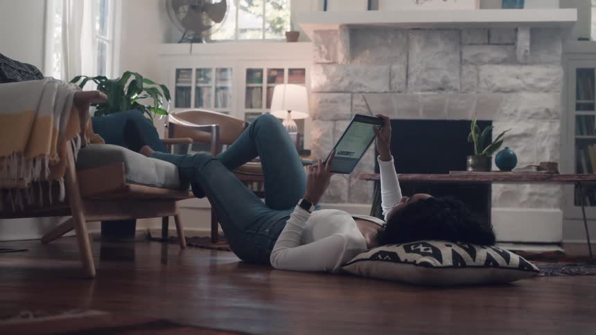 A woman lies on the floor with her feet on a living room chair scrolling and reading on her tablet
