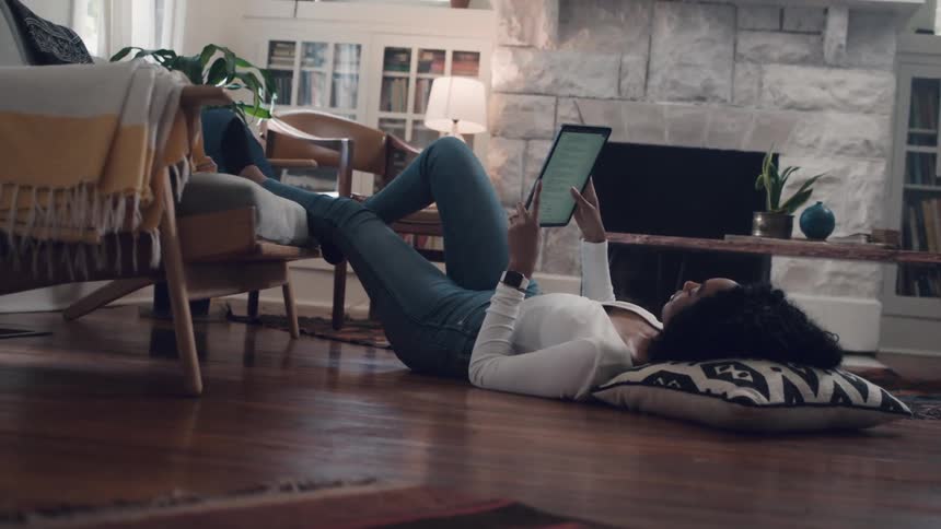 Black woman lying down on the floor uses a tablet in the living room at home