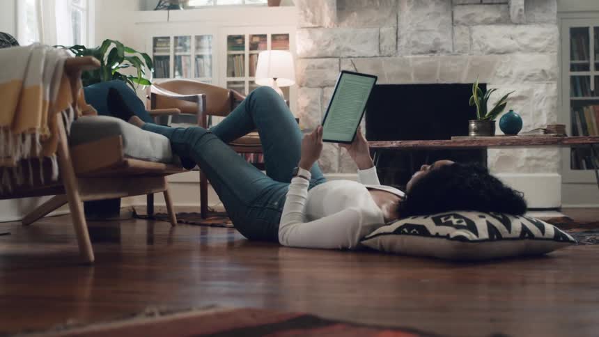 Woman lying down on the ground looks at her tablet in the living room at home