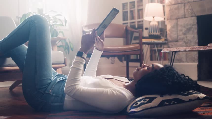 A woman laying on the floor with her head on a pillow and her foot on a couch while she is watching a tablet