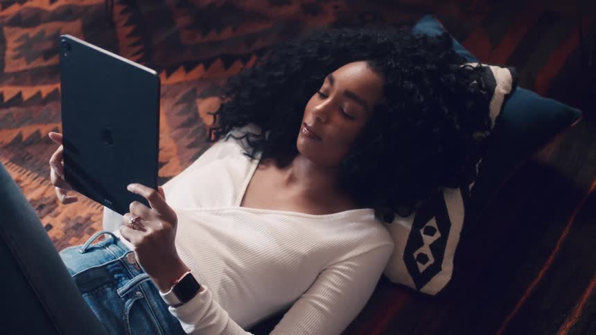 Black woman lying down on the floor uses a tablet in the living room at home