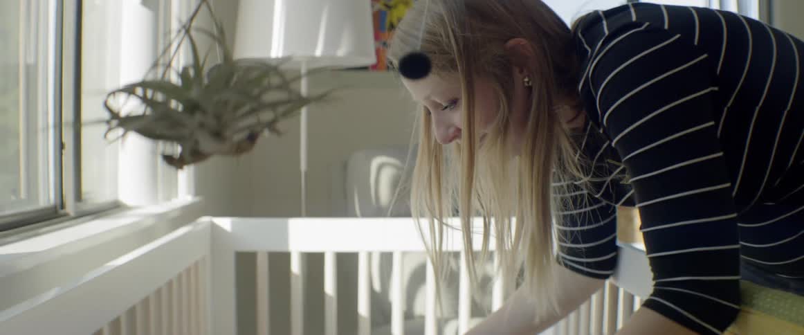 A mom adjust the mobile over her baby daughters crib then reaches in and holds her hands