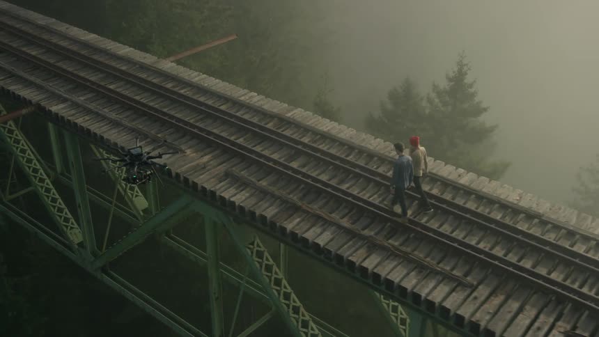 Two people walk across a railroad trestle bridge as a drone passes overhead