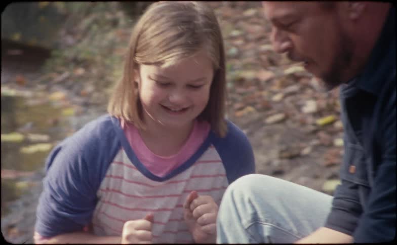 A young girl with her father and sister at a river picks up a worm and gets grossed out