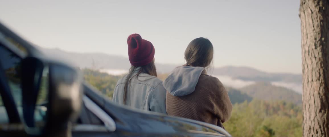 Two teenagers lean on a car and look out at the land and talk with one another