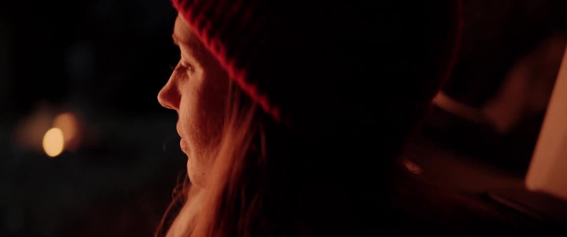 A woman in a beanie hat sits near a campfire at night