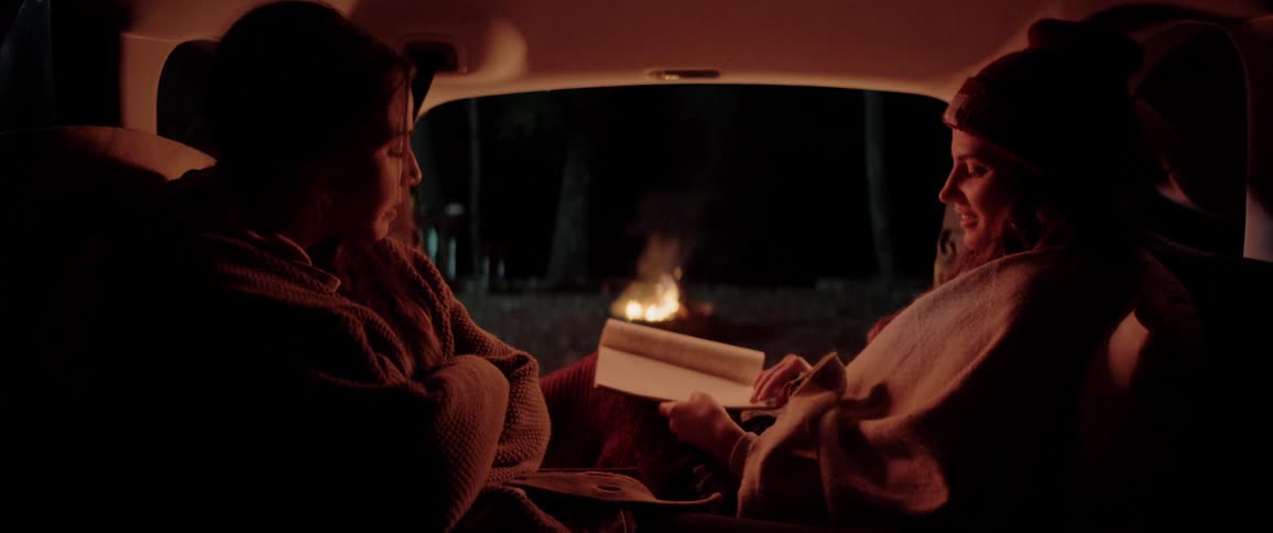Two young women inside a car talking and reading a book close to a camp fire at the forest