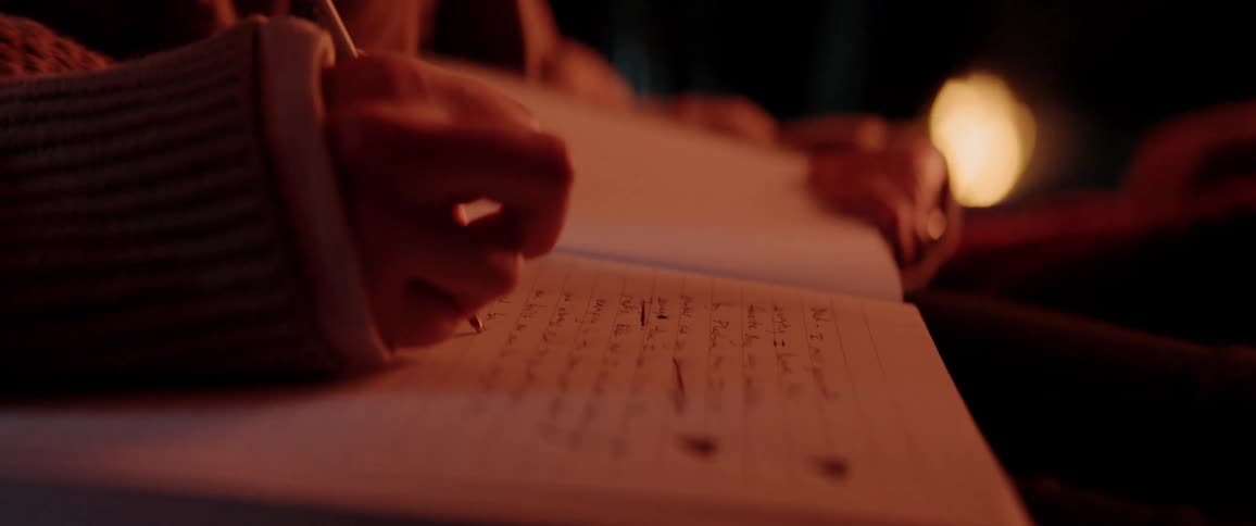 A person writes in a notebook sitting by a fire