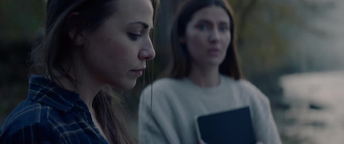 Two women stand on a river bank in a somber mood