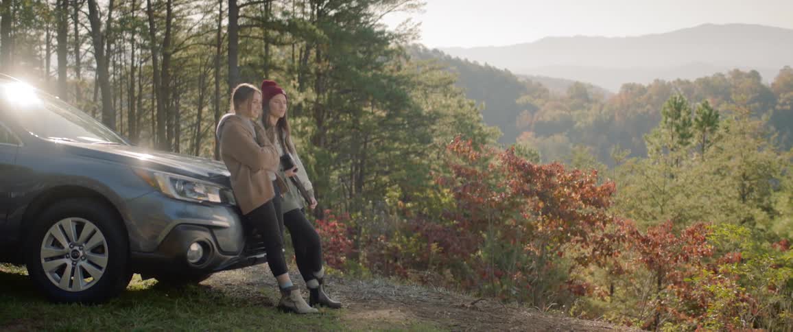 Two women lean against a car and watch the forest