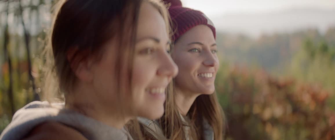 Two women smile and talk as they stand near the forest