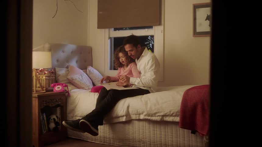 A father reads to his young daughter as they sit together on her twin bed