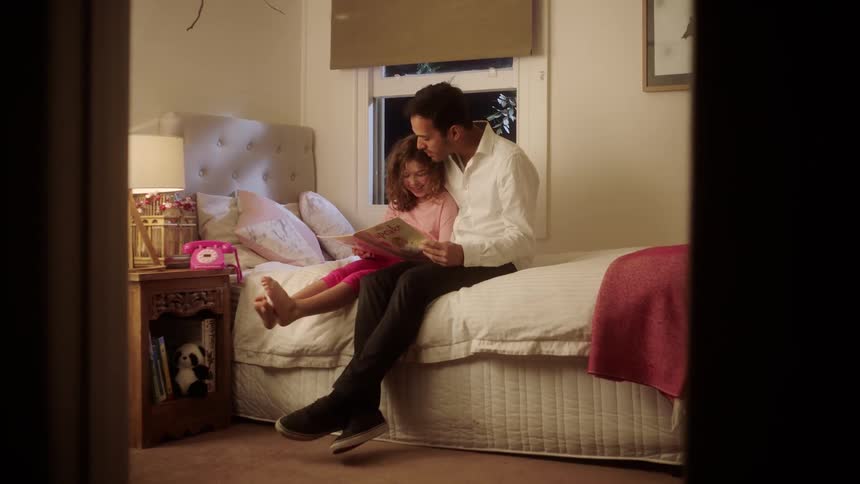 Father and daughter sitting at a bed while he reads a book for her