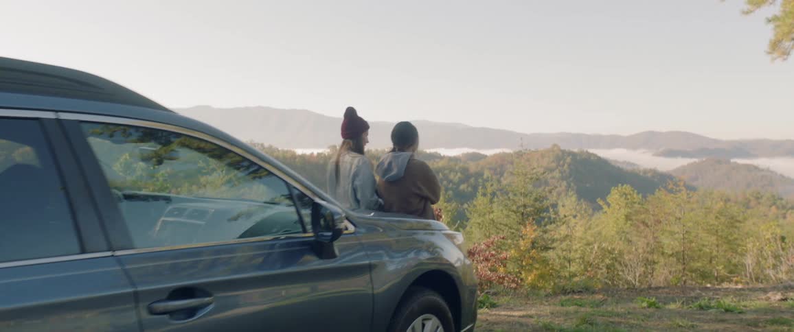 Man and woman lean against the front of a vehicle as they look out at the mountains