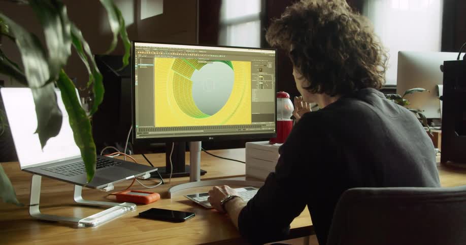 Male sitting at a desk in front of a computer monitor and a laptop while designing a 3d image