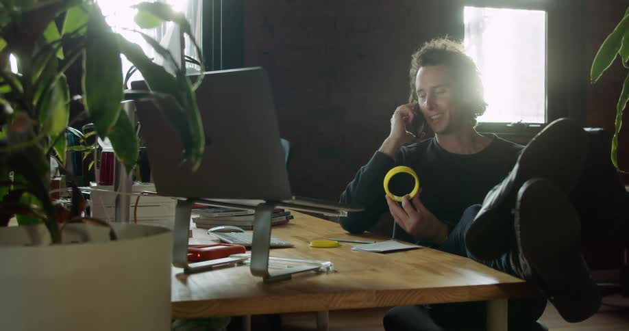 A man sits at his desk with his feet up talking on the phone while holding a yellow plastic ring
