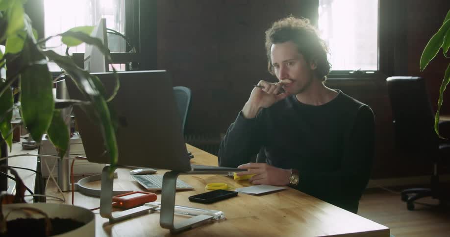 A man sits at his desk thinking while looking at his computer and a piece of yellow plastic