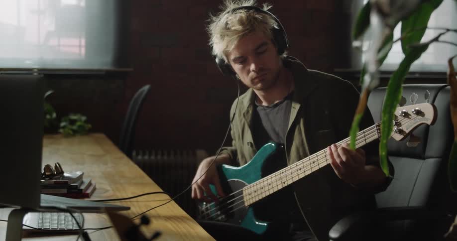 Man sits in chair in office wearing headphones and playing guitar while viewing monitor