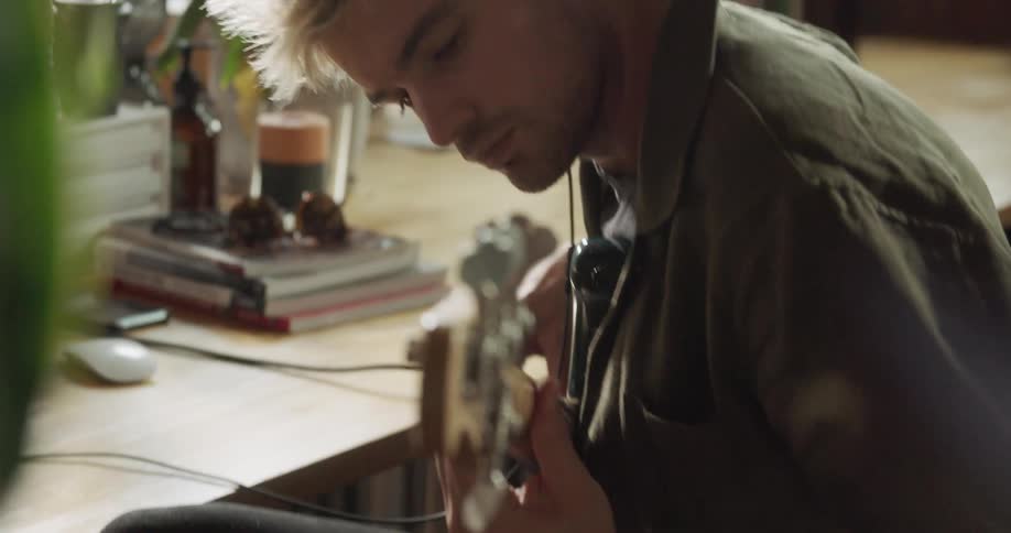 A man sits at a desk playing a guitar and wearing headphones