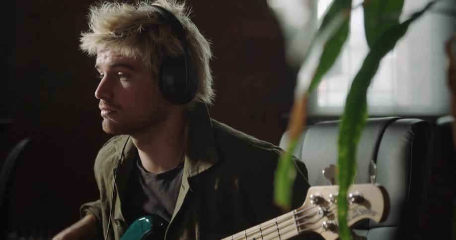 A man plays an electric guitar sitting at a desk wearing headphones