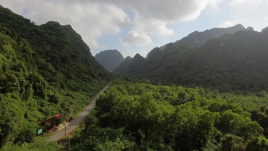 Road cuts through forest and mountains past buildings under cloudy sky