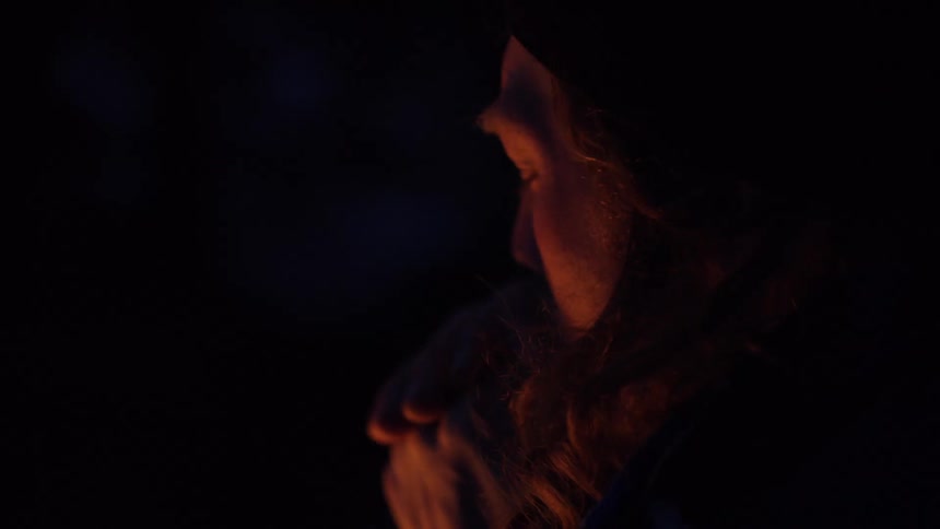 Firelight lights mans face as he warms hands outdoors among snow covered trees