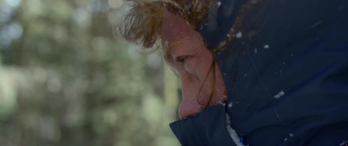 A man in a winter coat is standing in a forest while the wind blows snow flurries into his face