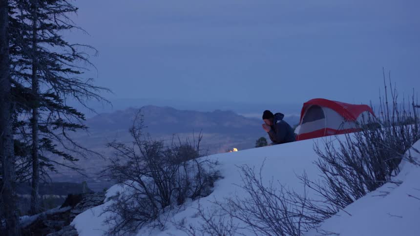 Man sitting in front of the campfire warms his hands at dusk