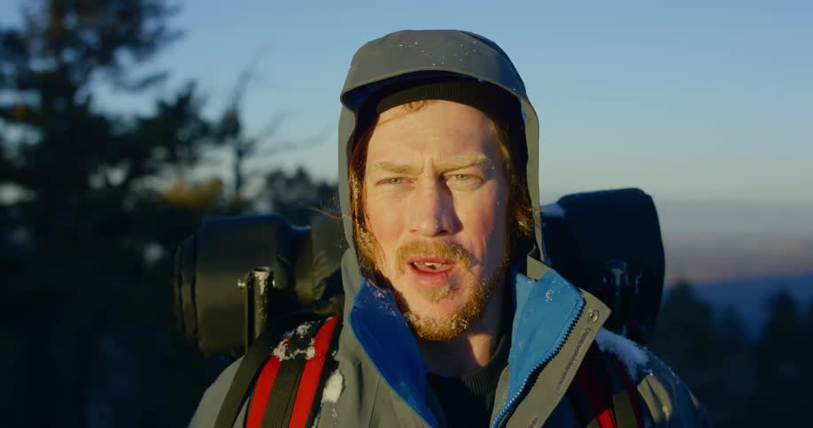 A man in a heavy jacket with camping gear breathes heavily as if just finishing a long hike