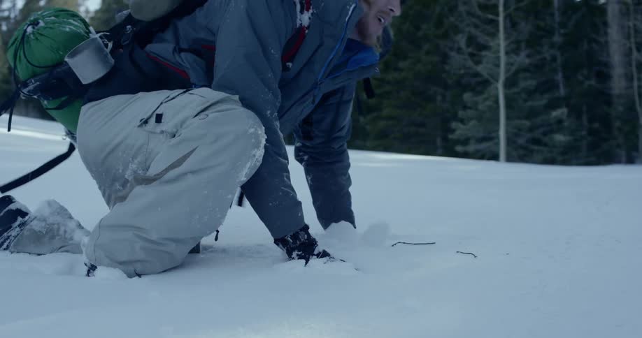 A man with cargo pants an overcoat and gloves getting up on his feet in the snow
