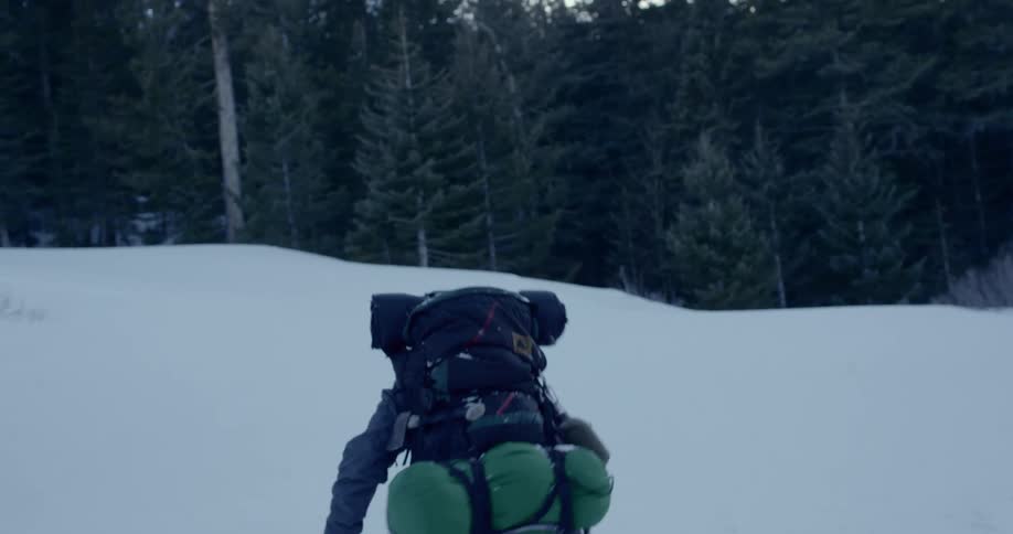 A person walks through deep snow towards a treeline wearing a heavy backpack and camping gear