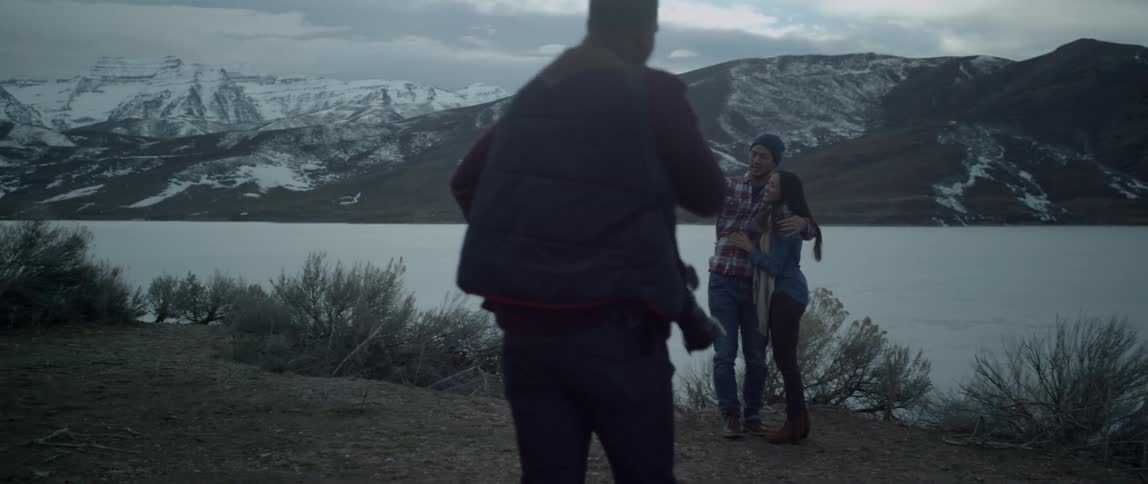 A man holding a camera walks up to two people posing by a mountain