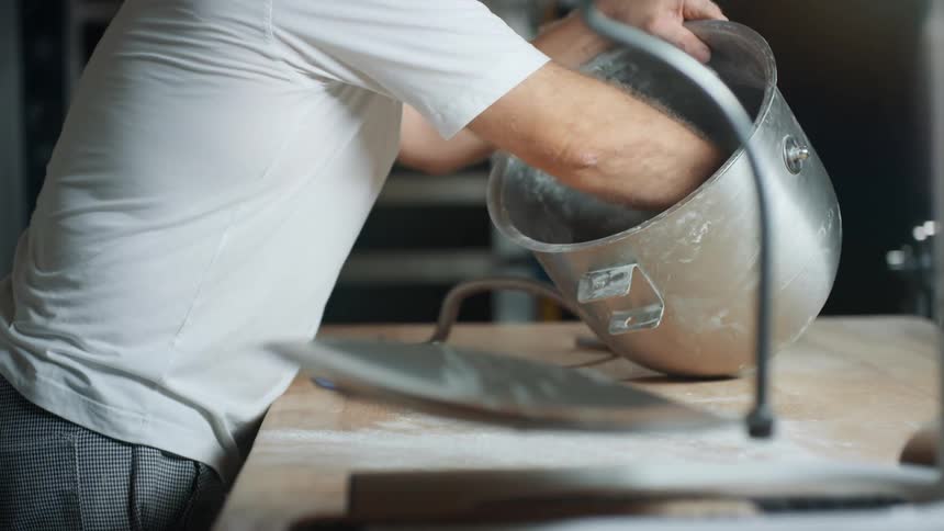 A man in a commercial kitchen area is collecting dough from a large mixing bowl