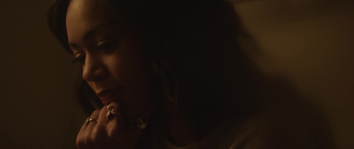 A woman puts her hand to her chin in a dark room