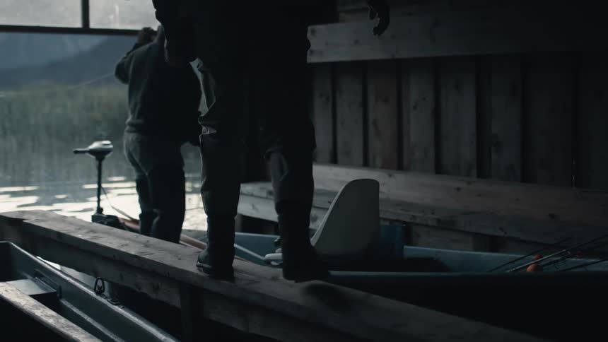 Two men get into a boat inside of a boat house that is equipped with fishing rods and a net