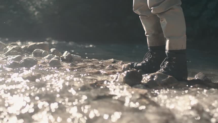 A man wearing khakis and black boots stands on rocks in a shallow river