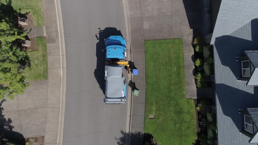 A garbage truck picks up a trash can outside a house in a neighborhood with well manicured lawns