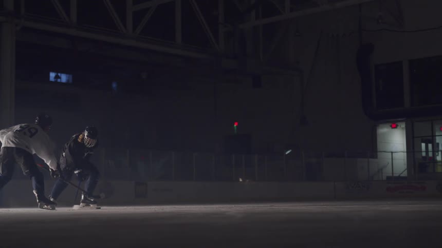 Two people practice ice hockey together in a dark rink