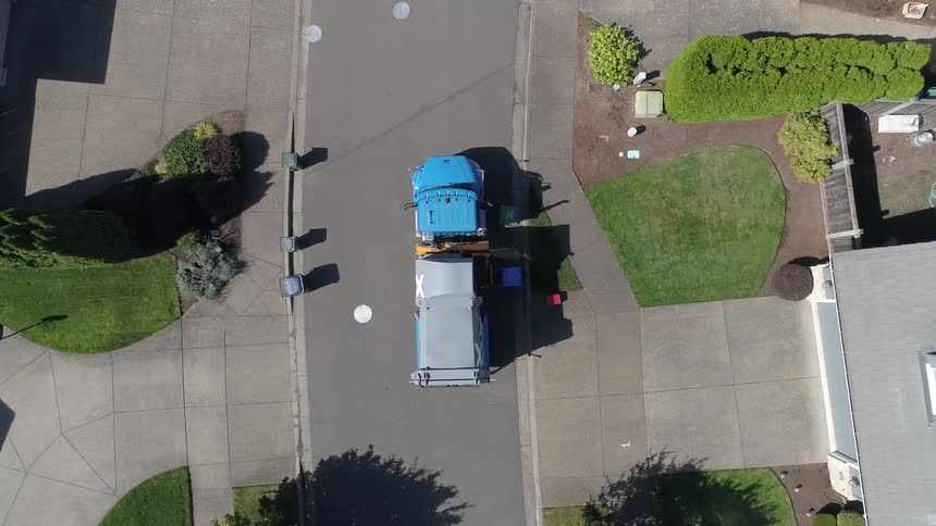 A trash pick up truck stops to pick up a trash can in front of a home and then drives away