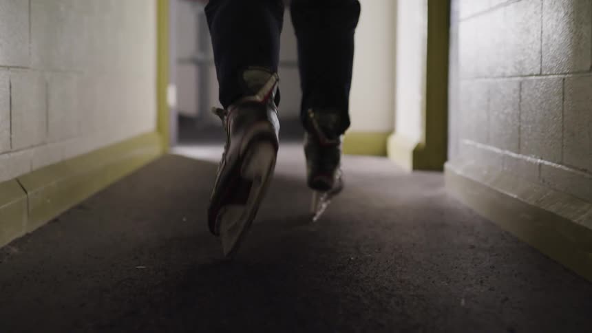 A man wearing skates and carrying a hockey stick walks down a hall