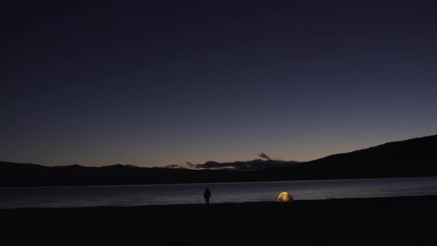 A person walks towards their lit up tent next to a river at dusk