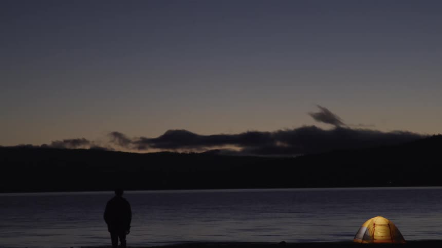 A person walks from a lake toward a lighted tent at night